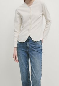 White, long-sleeve, button-up cardigan with a rounded hemline and textured fabric, paired with blue straight-leg jeans.
