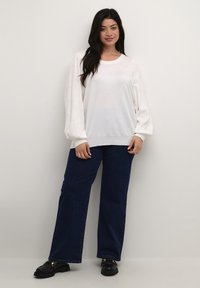 Vit tröja med texturerade puffärmar och rund halsringning, i kombination med mörkblå vida jeans och svarta loafers.