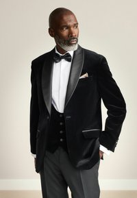 Zwarte fluwelen smokingjasje met satijnen revers, gecombineerd met een wit shirt en een zwarte vlindertie. Heeft een pochet met een gemusterde accent.