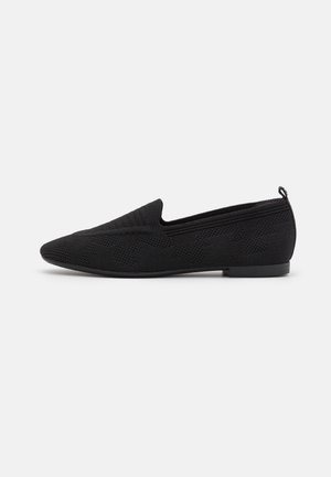 Sort strikket slip-on flad sko med spids tå, lav hæl og træktab på bagsiden mod hvid baggrund.