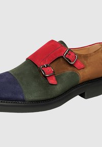 Mehrfarbiger Wildlederschuh mit einem marineblauen Zehenschutz, grünem Obermaterial, braunen Akzenten und einem roten Schnallenriemen mit silbernem Beschlag. Flache schwarze Sohle.