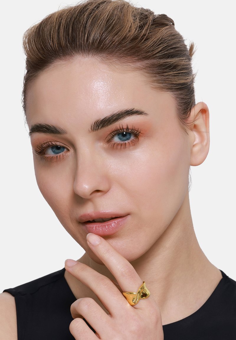 Gouden ring met een draaiend ontwerp, gedragen aan de vinger. Model raakt haar kin aan en toont gladde huid en blauwe ogen. Eenvoudig zwart topje.