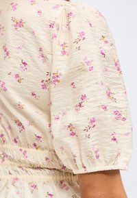 Μπεζ ύφασμα με μικρά ροζ και μωβ λουλουδάτα σχέδια, με φουσκωτό μανίκι με ελαστικό μανσέτα και λεπτομέρεια μαζεμένης μέσης.