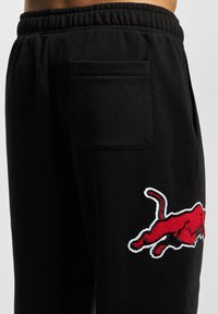 Svarta sweatpants gjorda av mjukt tyg, med ett elastiskt midjeband och en bakficka. En röd broderad logotyp finns på vänster lår.