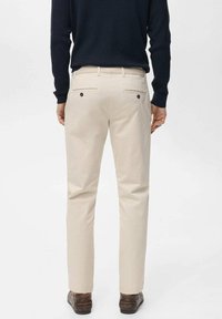 Light beige trousers with a smooth texture, featuring two back pockets with black buttons. Worn with a dark navy sweater and brown shoes.
