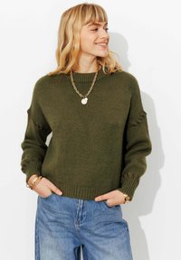 Pull en tricot vert olive avec un col rond, des poignets côtelés et des détails sur les manches. Porté avec un jean denim bleu ample.