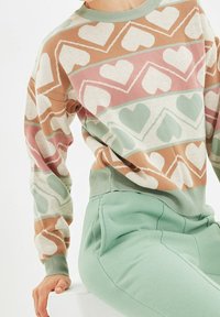 Person med en pastellfärgad tröja med hjärt-mönster i beige, rosa och grön, ihop med ljusgröna högmidjade byxor, som sitter på en vit yta.