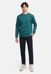 Felpa color teal in misto cotone con scollatura rotonda, polsini e orlo a coste. Abbinata a pantaloni neri e sneakers beige.
