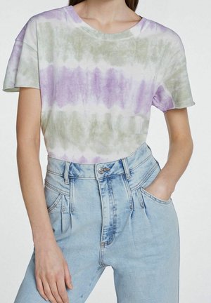 Femme portant un t-shirt tie-dye à manches courtes violet et vert, rentré dans un jean taille haute bleu clair avec des plis à l'avant.