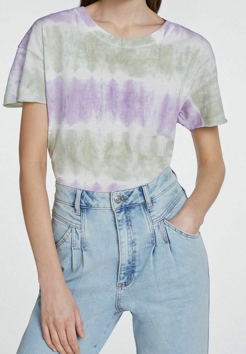 Femme portant un t-shirt tie-dye à manches courtes violet et vert, rentré dans un jean taille haute bleu clair avec des plis à l'avant.