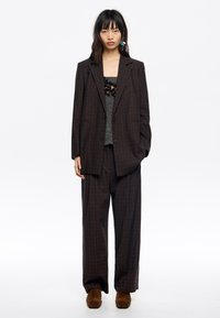 Completo a quadri marrone con giacca dal taglio morbido e pantaloni a gamba larga. Sovrapposto a un top grigio testurizzato, abbinato a scarpe in suede marrone.