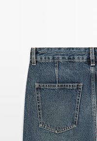 Tył niebieskich dżinsów z denimu ukazujący pasek, szlufki na pasek oraz jedną tylną kieszeń z widocznymi szwami na białym tle.