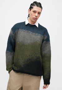 Πλεκτό πουλόβερ με οριζόντια διαβάθμιση σε αποχρώσεις βαθύ μπλε, γκρι, πράσινου και μαύρου. Με στρογγυλή λαιμόκοψη, μακριά μανίκια και απαλό ύφασμα. Φοριέται πάνω από λευκό πουκάμισο με γιακά.