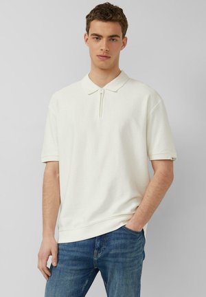 Junger Mann mit kurzen lockigen Haaren, der ein cremeweißes Poloshirt mit kurzem Arm und Reißverschluss am Kragen sowie eine blaue Jeans trägt, vor einem schlichten Hintergrund steht.