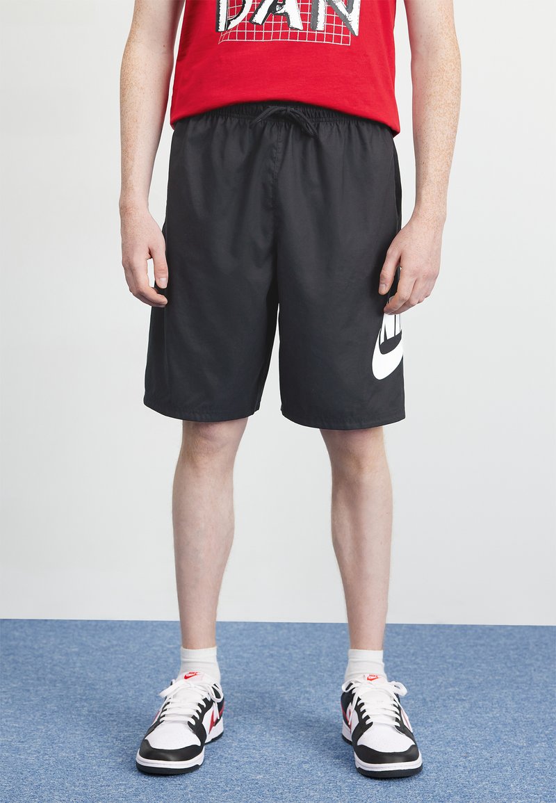 Black athletic shorts with elastic waistband and side pockets, featuring a large white Nike logo on the left side. Worn with white and black sneakers.