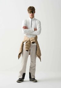 Haut blanc à manches longues avec fermeture éclair, veste matelassée beige nouée autour de la taille, pantalon beige et chaussures avec accents noirs. Lunettes de soleil argentées.