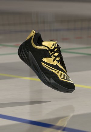 Baskets noires et dorées avec des accents dorés distinctifs flottant au-dessus du sol d'une salle de sport avec des lignes de terrain colorées.