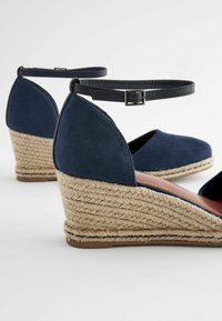 Sandali con tacco a cuneo in suede blu navy con cinturino alla caviglia, dotati di un tacco intrecciato in corda e foderati internamente in morbido marrone. Suola piatta per stabilità.