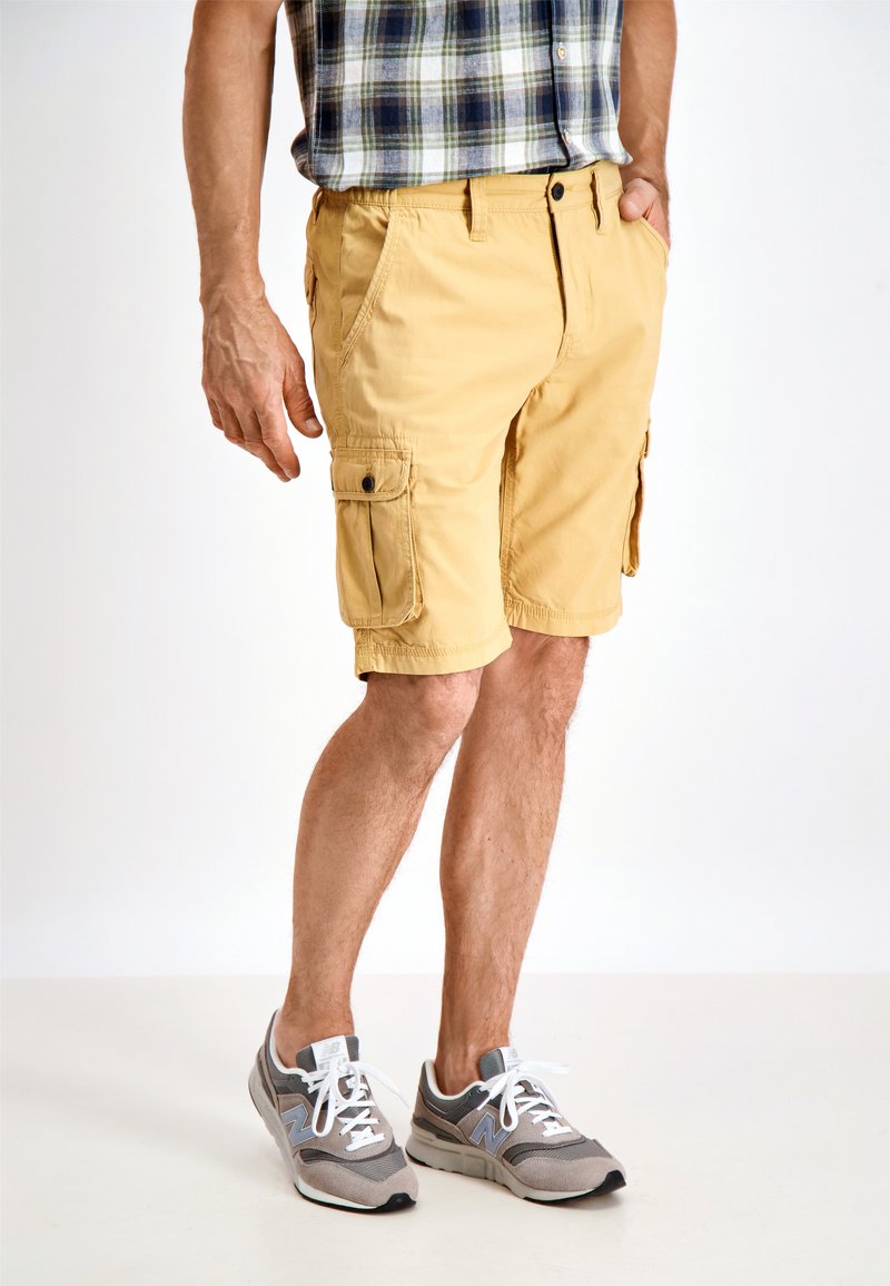 Pantaloni cargo gialli con tasche laterali, realizzati in tessuto leggero, caratterizzati da una vestibilità comoda e una texture semplice. Indossati con sneakers grigie.