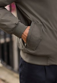 Graue Bomberjacke mit Reißverschlusstasche und elastischen Bündchen, aus glattem, wasserabweisendem Stoff und mit geripptem Saum. Detailaufnahme mit Hand in der Tasche.