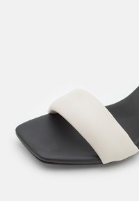 Sorte flade sandaler med firkantet tådesign, der har en bred hvid læderrem over toppen og en mat finish.