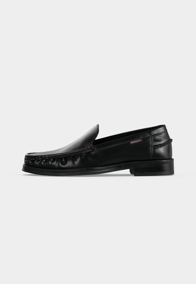 Mocassin noir en cuir à enfiler avec un petit talon et un détail moc toe cousu, présenté sur un fond uni gris clair.