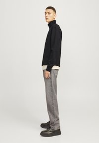 Black zip-up sweatshirt with high collar, gray denim jeans, and black sneakers. Model stands sideways against a neutral background.