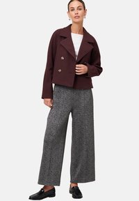 Femme portant un blouson bordeaux à double boutonnage, un haut blanc, un pantalon large gris et des mocassins noirs, se tenant devant un fond uni.