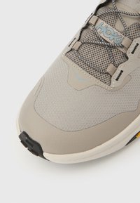 Ανοιχτό γκρι αθλητικό παπούτσι με επάνω μέρος από διχτυωτό υλικό, ενισχυμένη μύτη και μαύρη σόλα. Διαθέτει μπλε λεπτομέρειες και ελαστικό σύστημα κορδονιών.