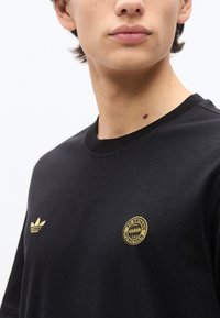 Νεαρό άτομο που φοράει μαύρο πουκάμισο με χρυσό λογότυπο Adidas αριστερά και έμβλημα της FC Bayern München στο δεξί στήθος.