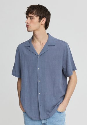 Młody mężczyzna w luźnej, krótkim rękawie, niebieskiej koszuli na guziki z kołnierzykiem, z rękami w kieszeniach, patrzący w lewo na jednolitym tle.