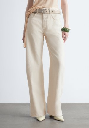 Pantalon beige à jambes larges porté avec une ceinture beige, chaussures crème pointues et bracelet vert au poignet gauche, debout sur un sol gris contre un mur blanc.