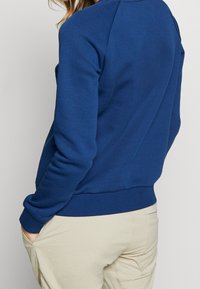 Modrá mikina se širokým výstřihem a žebrovanými manžetami. Vyrobena z měkké tkaniny, má volný střih a boční kapsy.