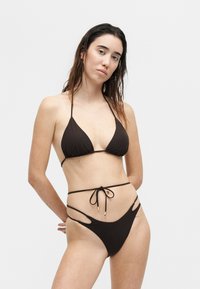 Mujer que lleva un bikini negro de tiras con top halter y braguitas de corte alto con recortes laterales, de pie frente a un fondo liso y claro.