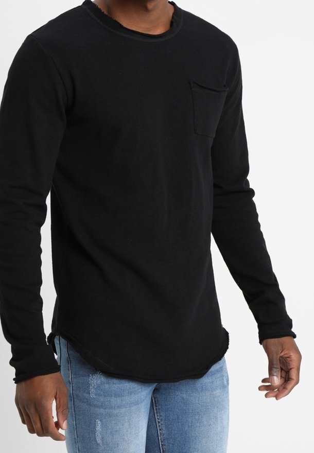 Person iført en sort langærmet skjorte med en brystlomme og lyseblå slidte jeans mod en hvid baggrund.