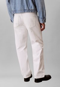 Άτομο που φοράει λευκό τζιν παντελόνι κανονικής γραμμής, μαύρα παπούτσια και ανοιχτό μπλε τζιν μπουφάν, εμφανιζόμενο από πίσω σε απλό φόντο.