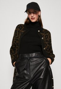 Luipaardprint cardigan in bruin en zwart, met knoopsluiting, gedragen over een zwarte coltrui. Gecombineerd met hoge zwarte leren broek.