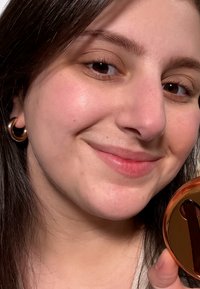 Nærbilde av et ansikt med glatt hud, iført gull hoop-øreringer, som holder et oransje, rundt speil med brun lokk. Naturlig belysning.