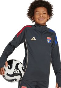 Veste de sport gris foncé avec des rayures rouges et bleues, des accents dorés et le logo de l'Olympique Lyonnais. Tient un ballon de football noir et blanc.