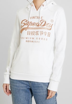 Sudadera con capucha blanca con texto metálico en oro rosa "SuperDry Vintage Premium Goods" y caracteres asiáticos, combinada con jeans azules sobre un fondo liso.