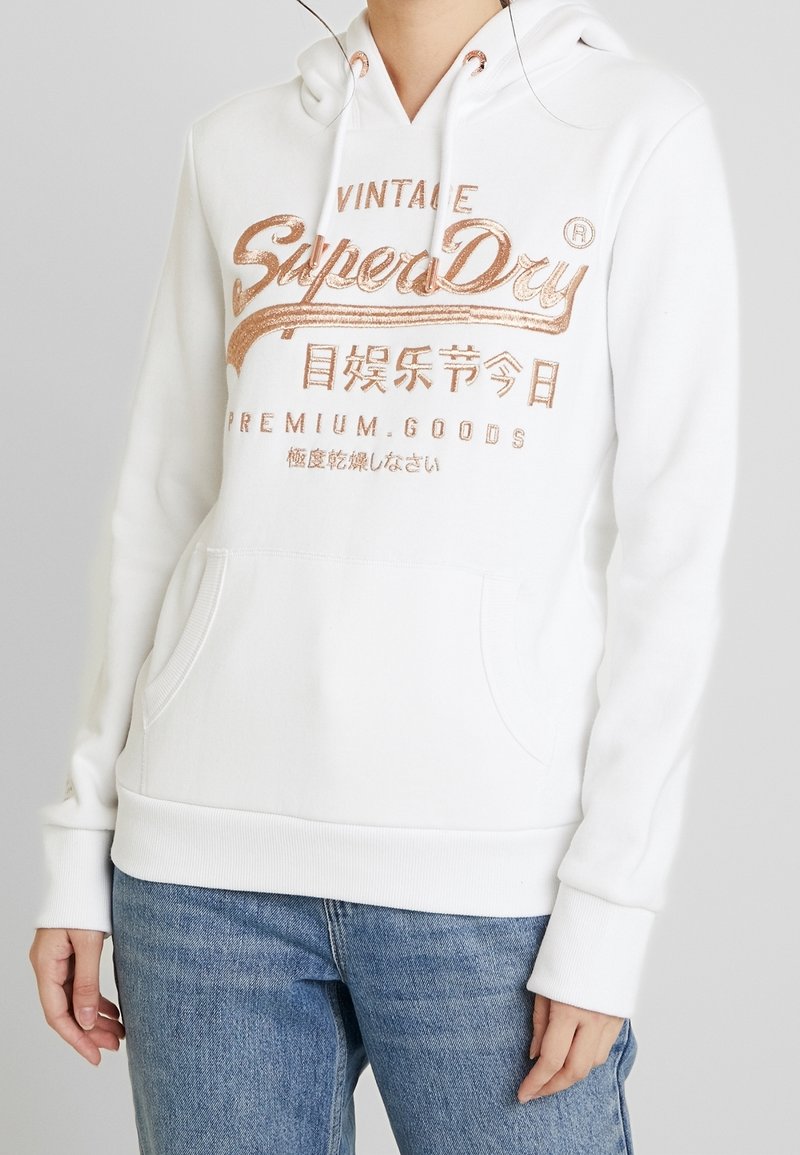 Witte hoodie met metalen roségouden tekst "SuperDry Vintage Premium Goods" en Aziatische karakters, gedragen met een blauwe spijkerbroek tegen een effen achtergrond.
