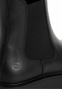 Gros plan sur une bottine Chelsea en cuir noir montrant un panneau latéral élastique et un logo circulaire embossé près de la semelle.