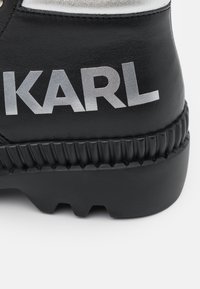 Tacón de zapato de cuero negro con suela gruesa acanalada y gran logo plateado "KARL" en el costado.