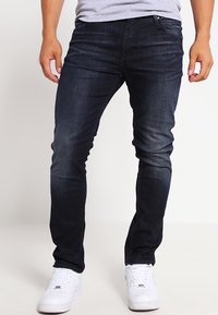 Tmavě modré džíny z denimu s úzkým střihem, klasickým pětiprvkovým designem kapes a minimálním vyblednutím. Nošeno s bílými teniskami.