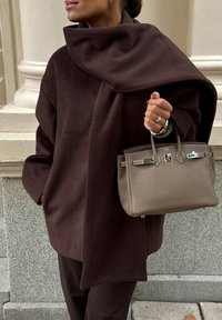 Pruun, ülemõõdulise suurusega fliiskurtaasi kõrge kaelusega, kantud koos väikese, heleda pruuni nahkkotiga, millel on hõbedased detailid.