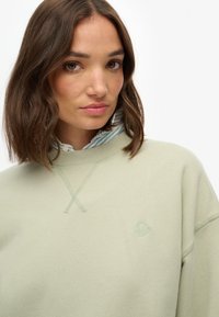 Felpa verde chiaro con una texture morbida, caratterizzata da un design a croce cucita sul colletto e un logo ricamato discreto sul petto.
