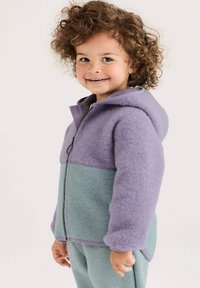 Lächelndes Kind mit lockigem Haar, das eine zweifarbige lila und türkisfarbene Fleecejacke mit Kapuze und passende Hosen trägt, vor einem hellen Hintergrund stehend.