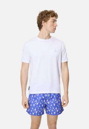 Joven con camisa blanca de manga corta con un pequeño logo de velero y pantalones cortos de baño azules con un patrón de veleros blancos, mirando hacia su derecha.