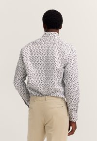 Mann mit gemustertem weißen Langarmshirt und beigen Hosen, von hinten vor einem schlichten hellen Hintergrund gezeigt.