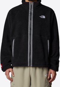 Svart fleecejacka med hög krage, dragkedjestängning, två sidofickor och grå sömmar. Har The North Face-logotypen på bröstet.
